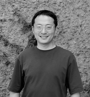 Black and white headshot of an Asian man with short dark hair and round glasses, smiling at the camera in front of a textured stone wall with vines.