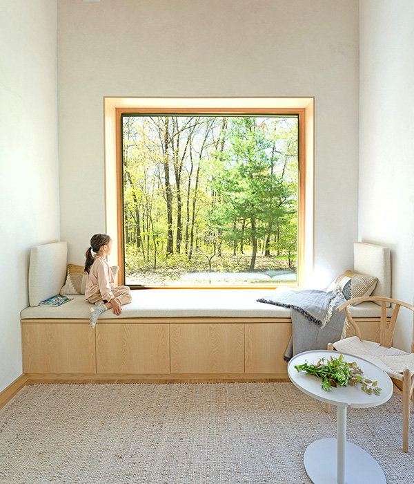 A bright interior space featuring a large square picture window that frames a sunlit green forest. Below the window is a wide, light-wood built-in window seat with an off-white cushion, throw pillows, and a gray fringed blanket. A young girl in light pink clothing sits on the left side of the seat, looking out at the trees. In the foreground, a textured natural fiber rug covers a light wood floor, accompanied by a small white round table holding leafy branches and a light wood wishbone chair.