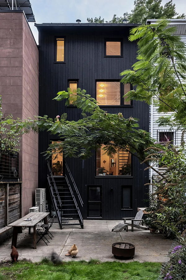 A rear view of a multi-story townhouse featuring a striking black vertical-plank facade with irregularly placed, warm-lit rectangular windows. A black metal staircase descends from a raised doorway to a concrete backyard patio furnished with a rustic wooden dining table, a lounge chair, and a fire pit. A leafy tree branch arches across the middle of the frame, partially obscuring the building. In the grassy foreground, two chickens are foraging. The black townhouse stands between a modern brown building on the left and a traditional white-siding building on the right.