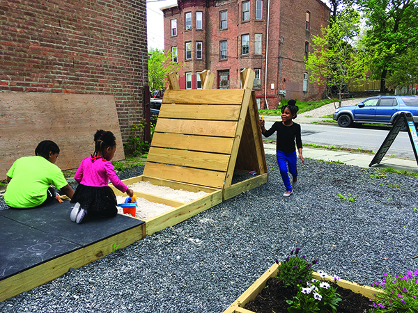 Three children playing in an urban pop-up park surfaced with grey gravel. Two children, wearing a green shirt and a pink shirt, are kneeling on a black rubber mat to play in a rectangular wooden sandbox. A third child, in a black top and blue pants, stands smiling beside an attached wooden A-frame play structure. A wooden planter box with flowers sits in the foreground, with a street, a parked blue SUV, and a multi-story brick building visible in the background.