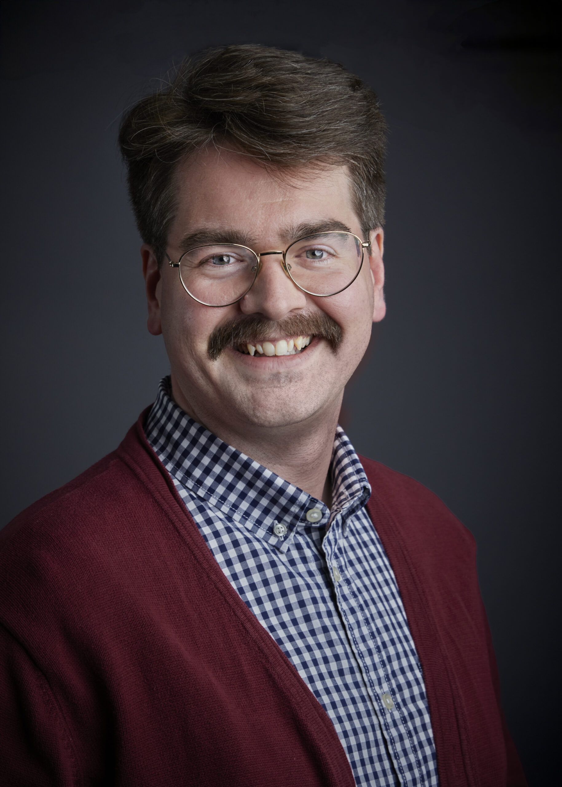 A man with dark hair and a neatly trimmed mustache smiles warmly at the camera. He wears round, wire-rimmed glasses and a blue and white checkered button-down shirt, layered under a red V-neck cardigan. The background is a solid, dark gray.