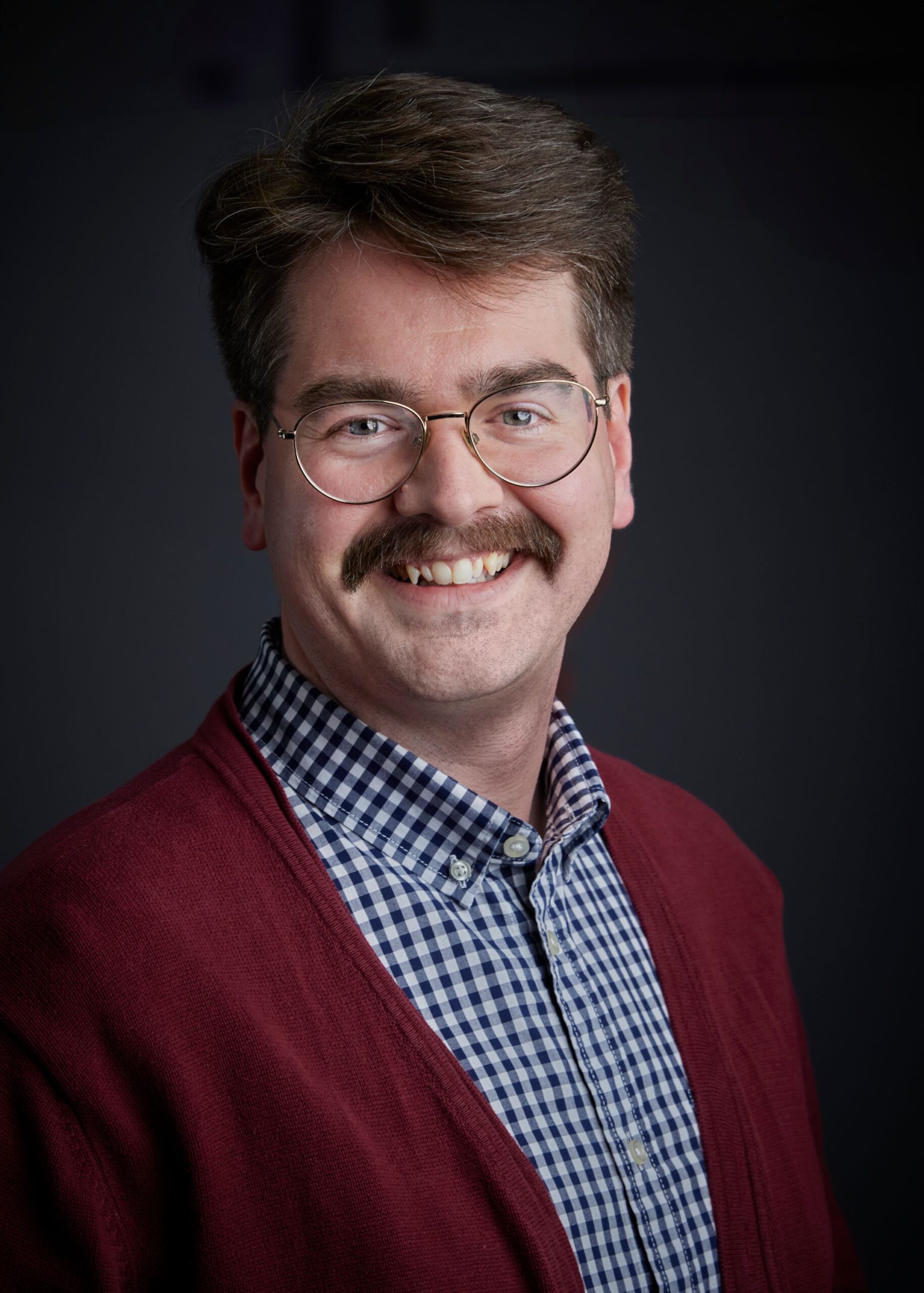 The portrait shows a man with fair skin, light brown hair styled with a side part, and a neatly groomed mustache. He is wearing round, gold-rimmed glasses and smiling, revealing his teeth. He is dressed in a blue and white checkered shirt, topped with a dark red cardigan. The background is a solid, dark gray.