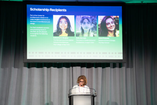 A woman speaks at a podium in front of a projection screen. 3 female scholarship winner headshots are projected on the screen behind her.