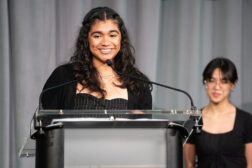 Photo of a young woman smiling and speaking at a podium. Another woman stands nearby in the background.