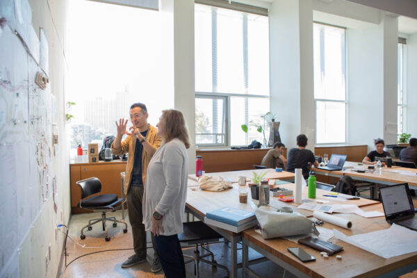 Prof. Zihao Zhang standing while conversing with a student during studio. Students are seated at a nearby table.