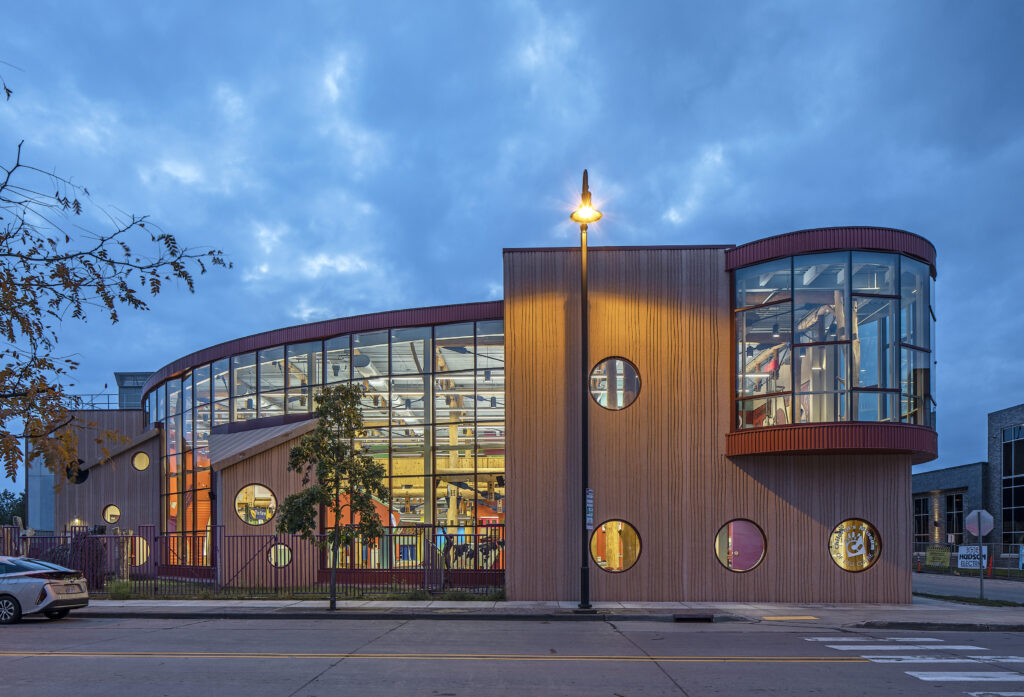 Facade of the Children's Museum of Eau-Claire with large glass windows and circular accents, warmly illuminated in the evening against a cloudy sky.