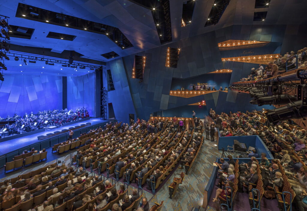 Interior of the Pablo Center at the Confluence Art Center, featuring a packed audience and an orchestra performing on a modern, angular stage illuminated in blue.