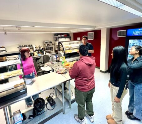 Three students wait in line to order at a coffee counter. The barista stands and smiles behind the counter.