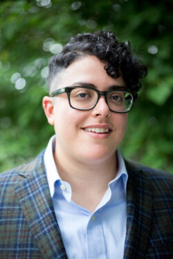 Image description: Aimi Hamraie, a nonbinary person with olive skin tone, short dark curly hair, and round black glasses, looks at the screen in front of leafy trees.