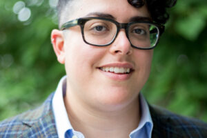 Image description: Aimi Hamraie, a nonbinary person with olive skin tone, short dark curly hair, and round black glasses, looks at the screen in front of leafy trees.