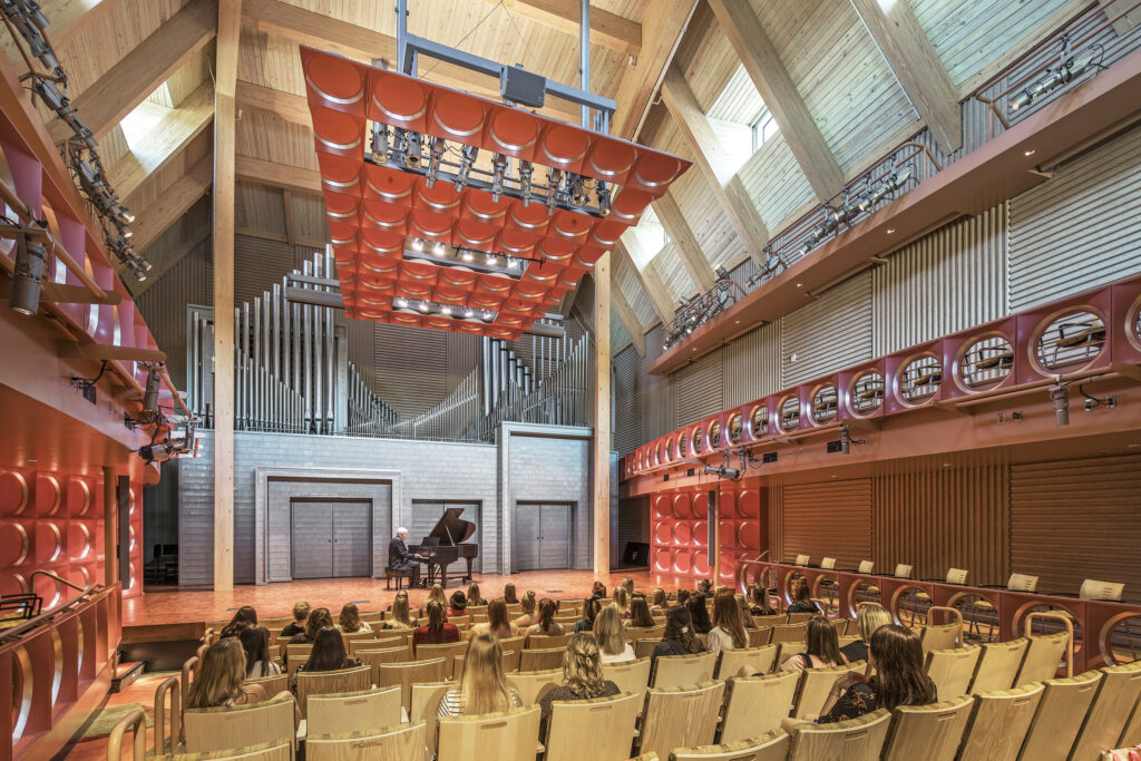 Interior of the South Dakota State University Performing Arts Center, showcasing a pianist performing on stage with an audience seated beneath a striking red acoustic ceiling and natural wood finishes.