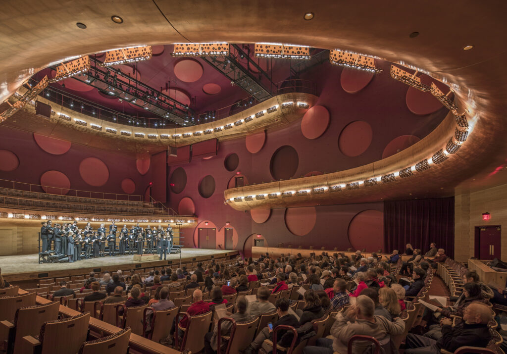 University Of Wisconsin Hamel Music, a concert hall with an audience seated before a choir performing on stage, surrounded by warm wood tones and circular acoustic panels on the walls.