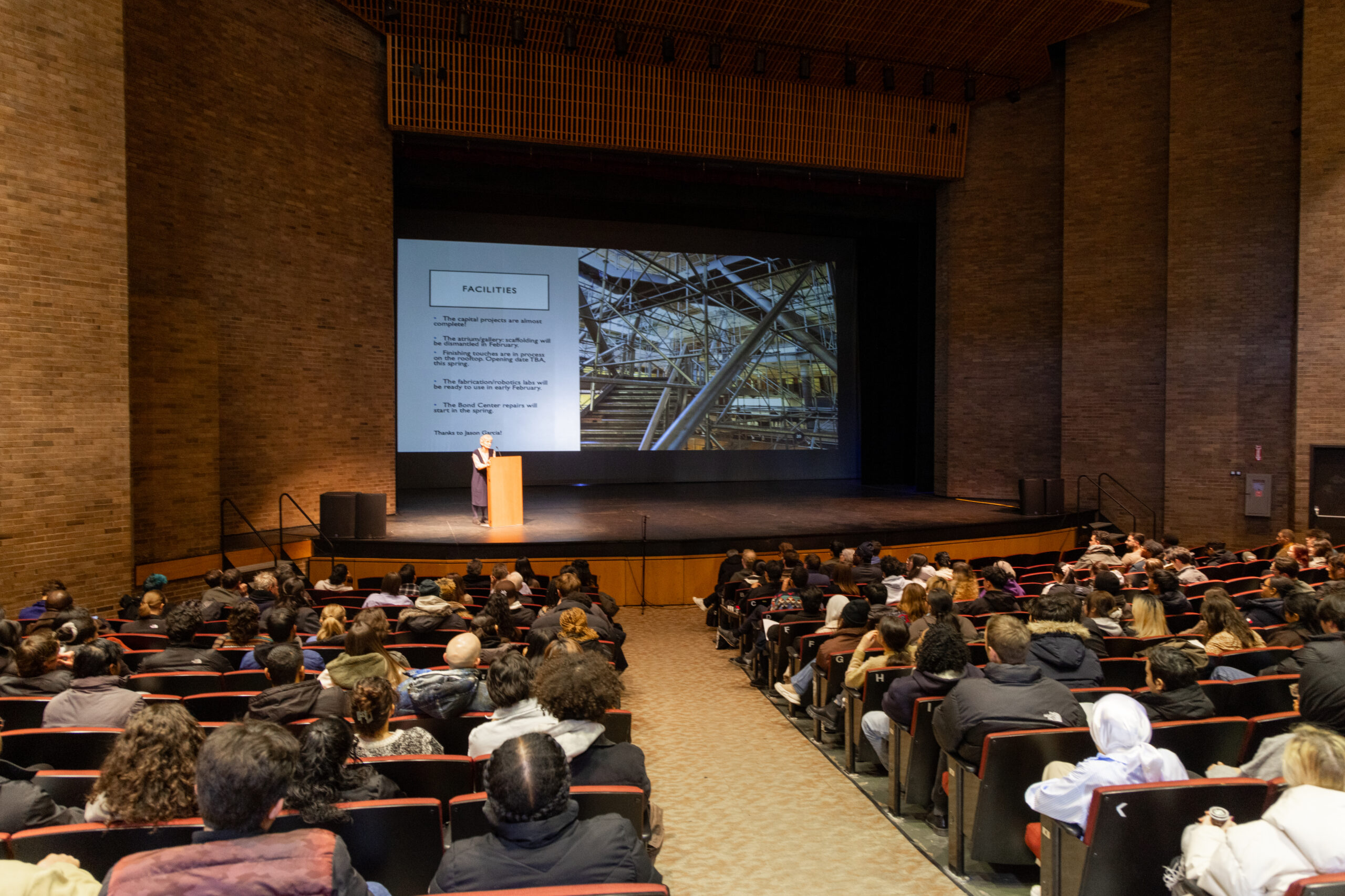 The Spitzer School of Architecture's Dean, Marta Gutman, stands at the podium in front of a crowd in an auditorium and delivers the Spring 2025 convocation address. A screen projects a presentation behind her.