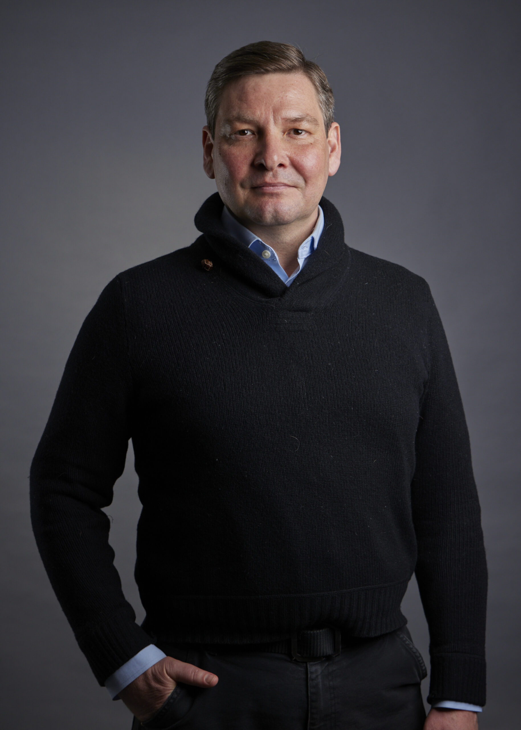 A portrait of a man with short brown hair, wearing a black shawl-neck sweater over a light blue collared shirt. He is looking directly at the camera with a neutral expression and has his right hand casually placed in his pocket. The background is a solid, dark gray.