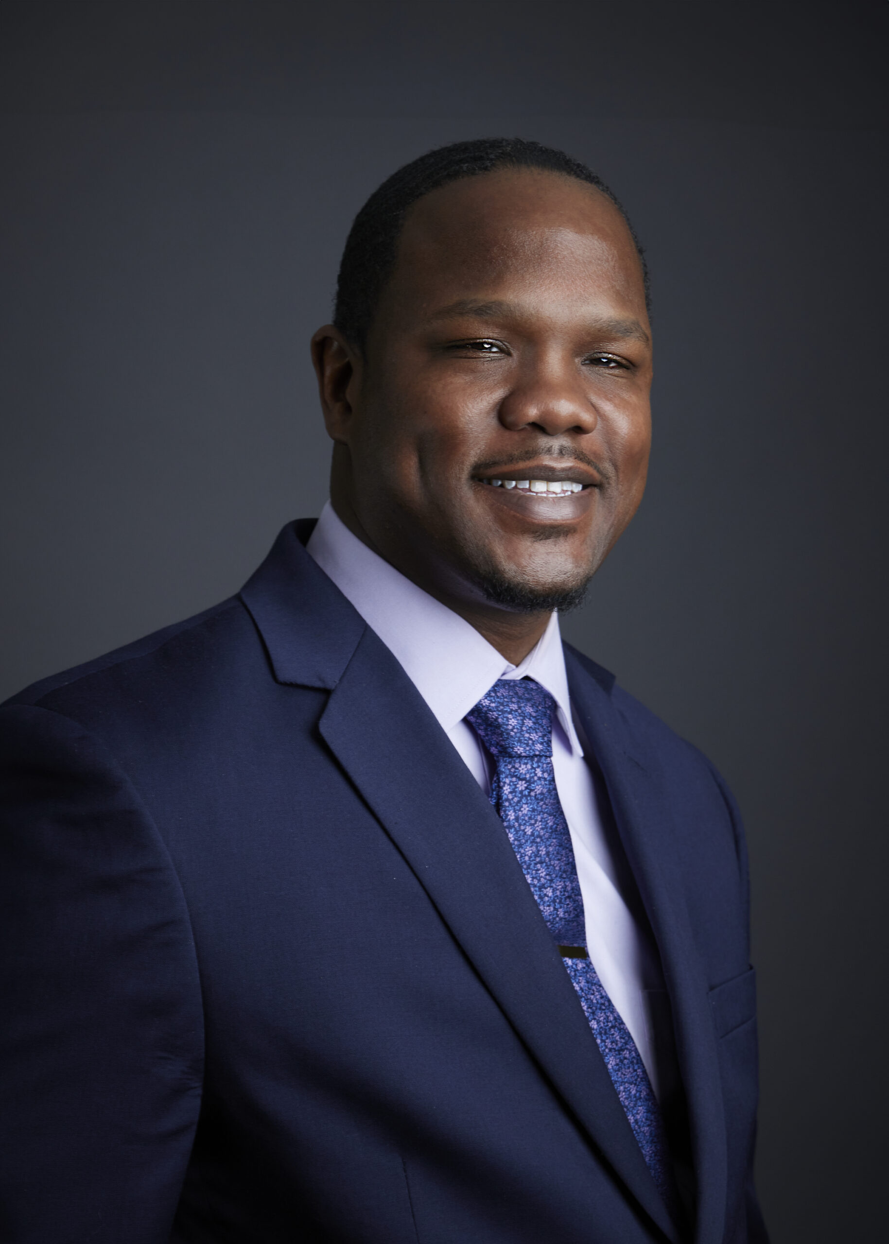 The portrait depicts a man with dark skin, short dark hair, and a neatly trimmed beard. He appears to be smiling. He is wearing a dark blue suit, a light-colored collared shirt, and a blue patterned tie. The background is a solid, dark gray.