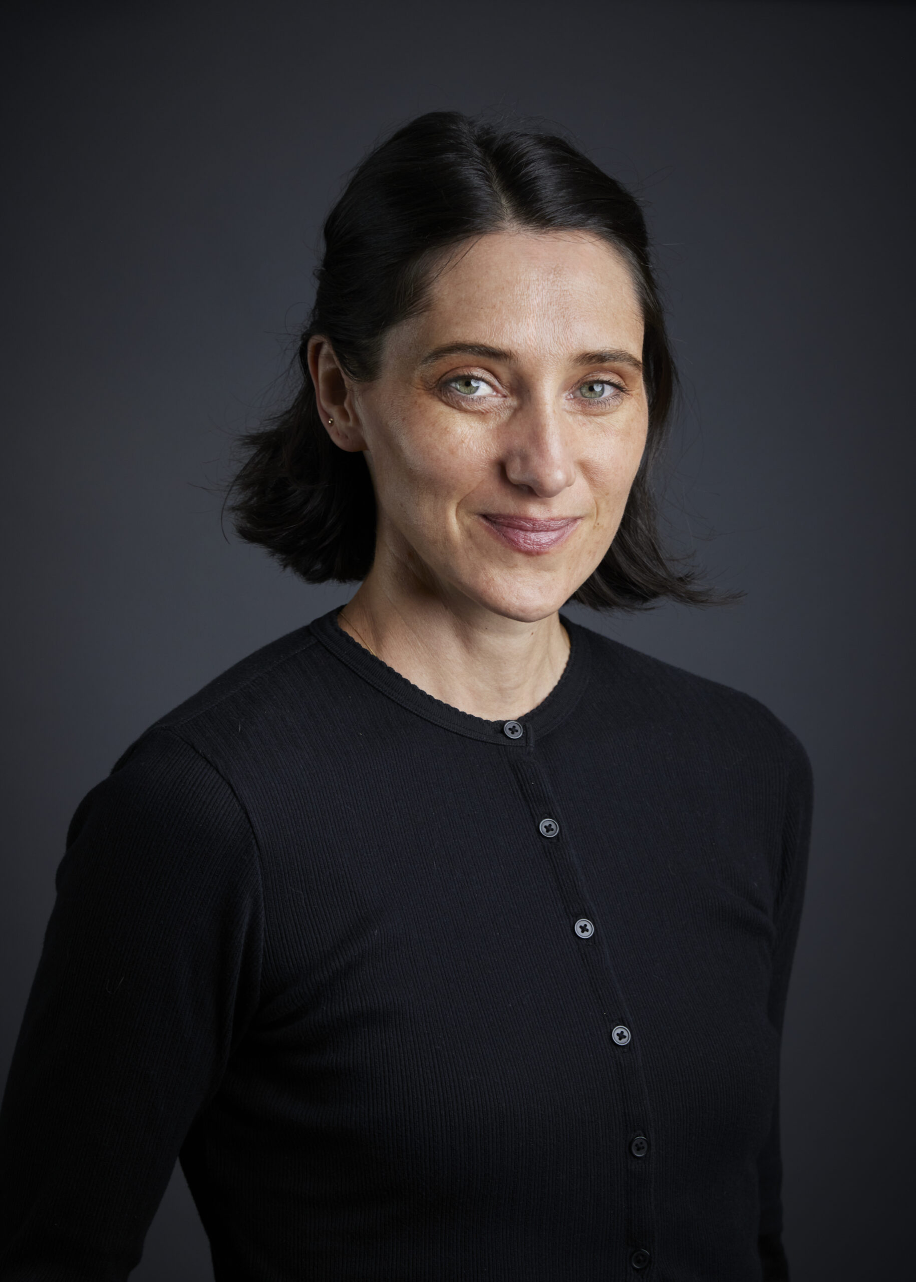 A portrait shot shows a woman with fair skin, dark brown hair parted slightly off-center and pulled back behind her ears, and light blue eyes looking directly at the camera with a soft, gentle smile. She has subtle freckles across her nose and cheeks. She is wearing a dark, possibly black, long-sleeved, button-up cardigan. The background is a solid, dark grey. The lighting is soft and even, highlighting her features without harsh shadows.