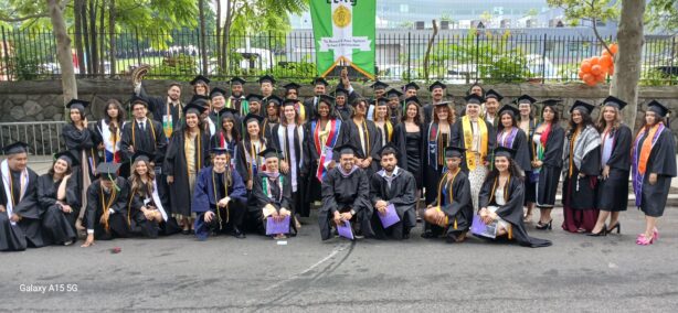 A large group of approximately 30 diverse graduates, dressed in black graduation caps and gowns with various colored stoles and sashes, pose outdoors on a paved area. In the background, behind a metal fence, is a street with some trees and buildings, and a green banner that reads 