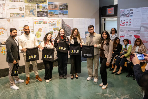 An indoor scene shows a group of seven adults, mostly standing, holding up black tote bags that appear to have a yellow logo on them, and one specifically saying 