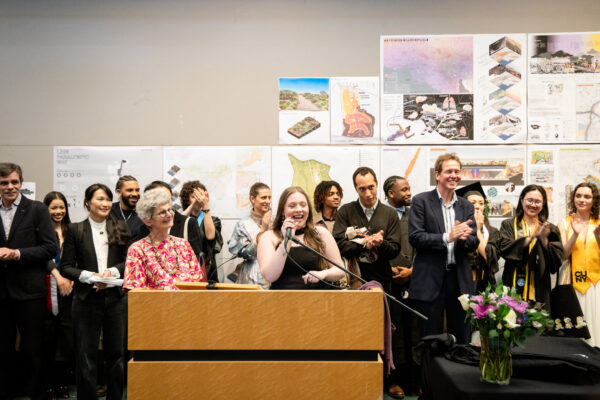 An indoor event featuring a large group of people gathered around a wooden podium. A young woman with long hair is speaking into a microphone at the podium, smiling broadly. To her left, an older woman in a floral top is also smiling, and behind them, a diverse group of individuals, some wearing graduation caps and gowns, are watching and smiling. To the right of the podium, a man in a suit is clapping, and a woman in a graduation cap and gown is also clapping. The background features display boards with various maps and architectural designs. A floral arrangement with purple and white flowers is visible on a table to the right. The atmosphere appears to be celebratory and engaging.