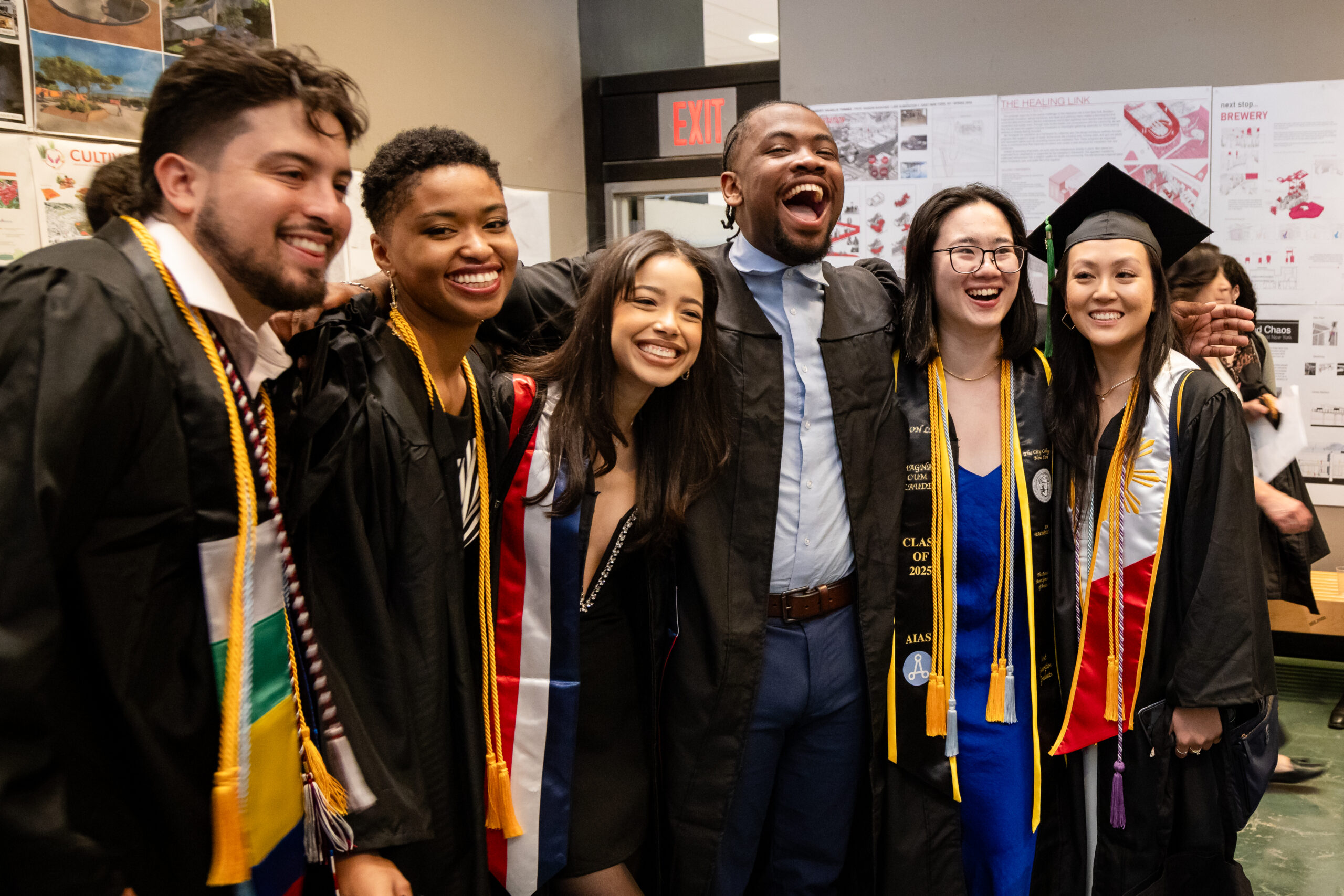 A joyful group of six young adults, dressed in graduation caps and gowns with various colorful stoles, are posing closely together indoors. They are all smiling and laughing, looking towards the camera, with one person in the center appearing to be mid-laugh with their mouth open. The background features white walls with what appear to be architectural or design project boards displayed, and a red