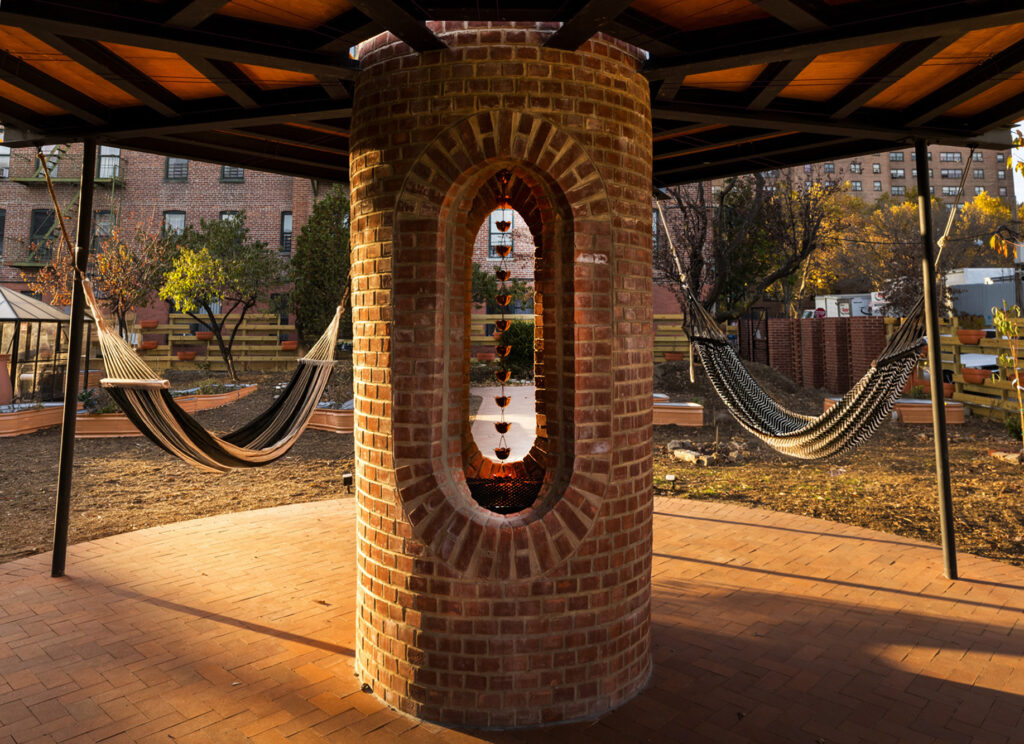 A brick structure with two large, arched openings stands under a wooden roof. Two striped hammocks hang from the structure's metal supports. The ground is paved with brick, and the background shows a community garden and apartment buildings under a clear sky.