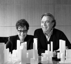 A black and white photograph of two people, a woman and a man, sitting behind a table covered with a large-scale architectural model of a city. The woman on the left has short, curly hair and glasses, and is smiling. The man on the right has wavy hair and is also smiling broadly.