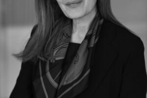 A black and white, vertical headshot of a woman with long, light-colored hair, smiling slightly and looking at the camera. She is wearing a dark jacket over a patterned scarf.