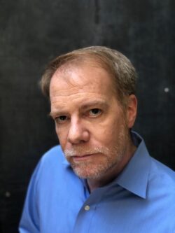 A portrait of a middle-aged man with a light-colored beard, wearing a blue collared shirt. He is looking directly at the camera with a serious expression against a dark, textured background.