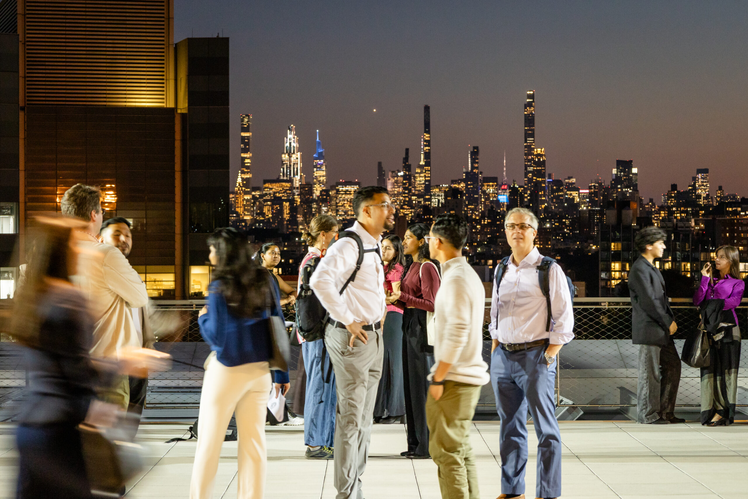 A group of diverse people are mingling and conversing on an outdoor terrace or rooftop at night. The brightly lit Manhattan skyline, including several tall skyscrapers, forms a dramatic backdrop to the networking event. Some individuals in the foreground are slightly blurred due to movement.