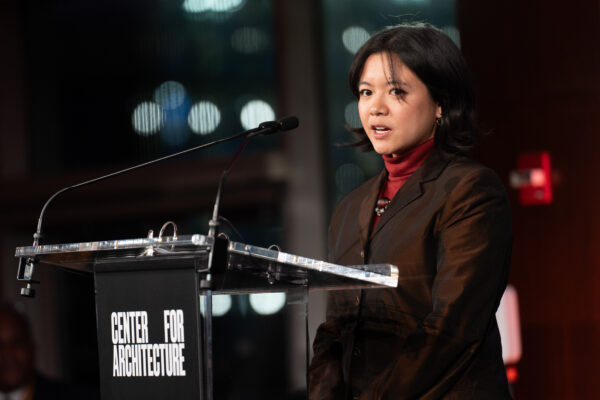 A medium close-up of a young woman, Justine Flora, speaking at a podium during an evening event. She is wearing a dark blazer over a red turtleneck and has dark, short hair. The podium is clear acrylic with a black base that reads 