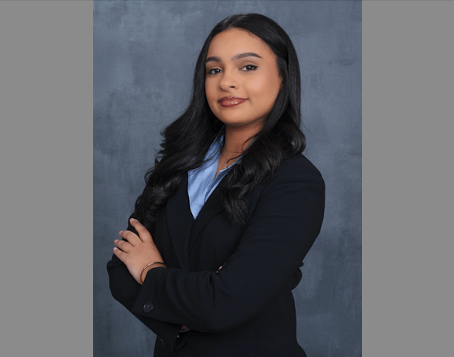 A professional headshot of a young woman with long, wavy dark hair and a warm, confident expression. She is posed at a slight angle with her arms crossed, looking toward the camera. She is wearing a dark navy blue or black blazer over a light blue collared shirt. The background is a textured, mottled grey-blue studio backdrop. The lighting is soft and even, highlighting her polished makeup and jewelry, which includes a gold bracelet and a delicate necklace.
