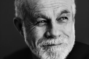 A black-and-white portrait of Carlos Moreno, a man with a short white beard and graying hair. He is looking slightly away from the camera with a thoughtful expression and a faint smile. The lighting creates high contrast, emphasizing his facial features against a dark, minimalist background.