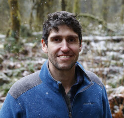A headshot of Ben Goldfarb, a man with short, dark curly hair and a light beard, smiling while wearing a blue fleece jacket. He is outdoors in a wooded area with light snow falling around him.