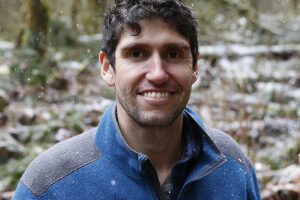 A headshot of Ben Goldfarb, a man with short, dark curly hair and a light beard, smiling while wearing a blue fleece jacket. He is outdoors in a wooded area with light snow falling around him.