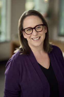 A headshot of Megan Nielsen Hegstad, a woman with shoulder-length brown hair and glasses with dark frames, smiling warmly while wearing a dark purple cardigan over a black top.