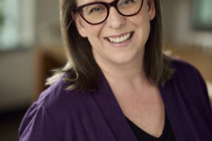 A headshot of Megan Nielsen Hegstad, a woman with shoulder-length brown hair and glasses with dark frames, smiling warmly while wearing a dark purple cardigan over a black top.
