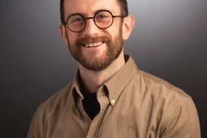 A headshot of a smiling man with a beard and round, black-rimmed glasses, wearing a tan corduroy button-down shirt over a black t-shirt against a dark grey gradient background.