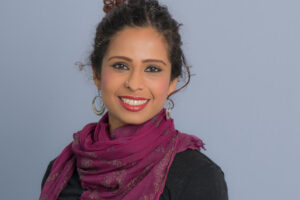 A headshot of Yamini Narayanan, a woman with dark hair styled in a bun, wearing a black top, a vibrant purple scarf with small patterns, and large silver hoop earrings, smiling against a light blue background.
