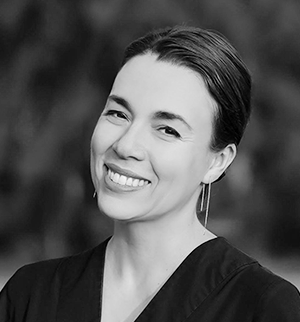 A black and white headshot of a smiling woman with her hair pulled back, wearing a dark V-neck top and long, thin drop earrings.