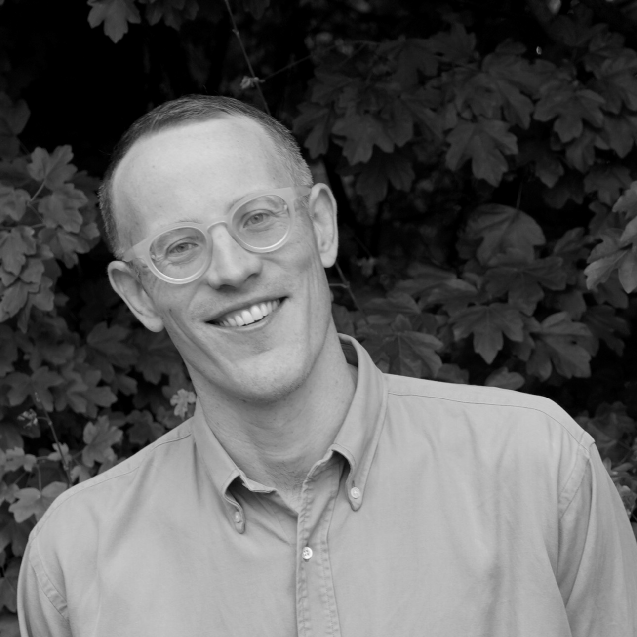 A black and white, square-format headshot of a smiling man with short hair and light-colored, round-framed glasses. He is wearing a button-down shirt and standing in front of a background of leafy foliage.