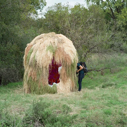 A large, rounded hut made of dried grass and hay stands in a grassy field surrounded by trees. A small opening in the front reveals a glimpse of a vibrant red and purple patterned interior. A person with a backpack stands to the right, looking toward the structure.