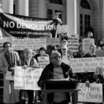A black-and-white photo of a protest on building steps against the demolition of public housing in Chelsea. A woman speaks at a lectern, backed by a diverse crowd holding signs and a central banner reading: "SAVE OUR HOMES: NO DEMOLITION - FULTON & ELLIOTT-CHELSEA HOUSES." Other visible signs demand, "My Home My Choice," "Preserve Retro Historic Buildings," and call out the negative impact of "Privatization."