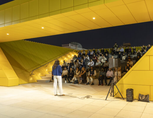 A wide-angle, eye-level outdoor shot at night shows a woman in a blue cardigan and white trousers standing on a light-colored stone patio, her back to the camera. She faces a large, diverse audience seated on stone bleachers that are part of a modern, bright yellow architectural structure with sharp angles and a large overhead overhang. The scene is illuminated by warm, artificial lights embedded in the structure. To the right of the woman, a black speaker on a tripod and a smaller amplifier on the ground are visible. The dark night sky is visible in the background.
