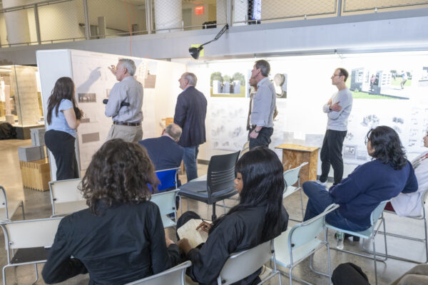 A small group of people sit on chairs or stand near white display boards on the wall, viewing images or presentations together.