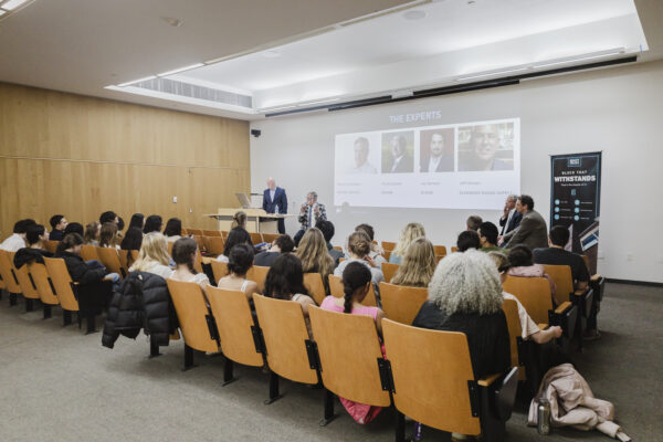A view from the back of a lecture hall showing an audience seated in wooden chairs, facing a presentation. At the front of the room, speakers are seated on a panel next to a large projection screen displaying headshots and names of the presenters.