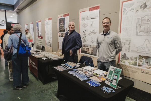 Two men stand behind a long, black-covered table at a career fair or exhibition. Behind them, several large posters display architectural drawings and project information. On the table, there are flyers, brochures, and lanyards.