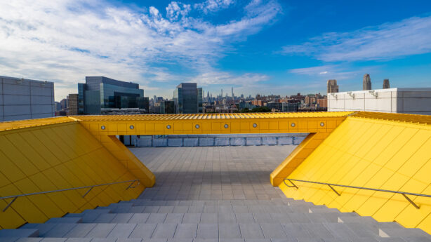 A high-angle, wide shot looking down a flight of gray stone bleacher stairs onto a flat, tiled rooftop terrace. The stairs are flanked by two bright yellow, angled architectural walls with metal handrails. A matching yellow horizontal beam spans across the top of the frame, creating a rectangular window-like view of the urban horizon. Beyond the terrace, a dense cityscape of various office buildings and skyscrapers stretches toward the horizon under a bright blue sky filled with wispy white clouds. The scene is captured in bright, natural daylight.