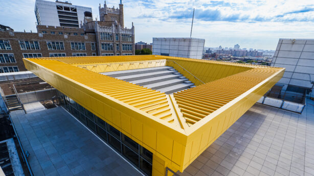 A high-angle, wide shot shows a modern rooftop terrace featuring a prominent, bright yellow rectangular structure that resembles an open-air amphitheater. This yellow feature has integrated gray stone bleacher seating that descends toward a tiled walkway. The surrounding rooftop is paved with large, light-colored square tiles and is bordered by safety railings and the glass walls of the building's upper floor. In the background, older brick academic-style buildings with gothic details contrast with the modern rooftop, all under a bright, partly cloudy sky. The urban landscape of the city extends toward the horizon.