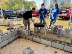Three individuals working at an outdoor construction site on a sunny day. A man in a black shirt leans over to scoop wet mortar from a tub elevated on concrete blocks, while two women in blue shirts look on. In the foreground, lower walls made of stacked concrete blocks with mortar are under construction on a dirt surface. Vehicles and trees are visible in the background.