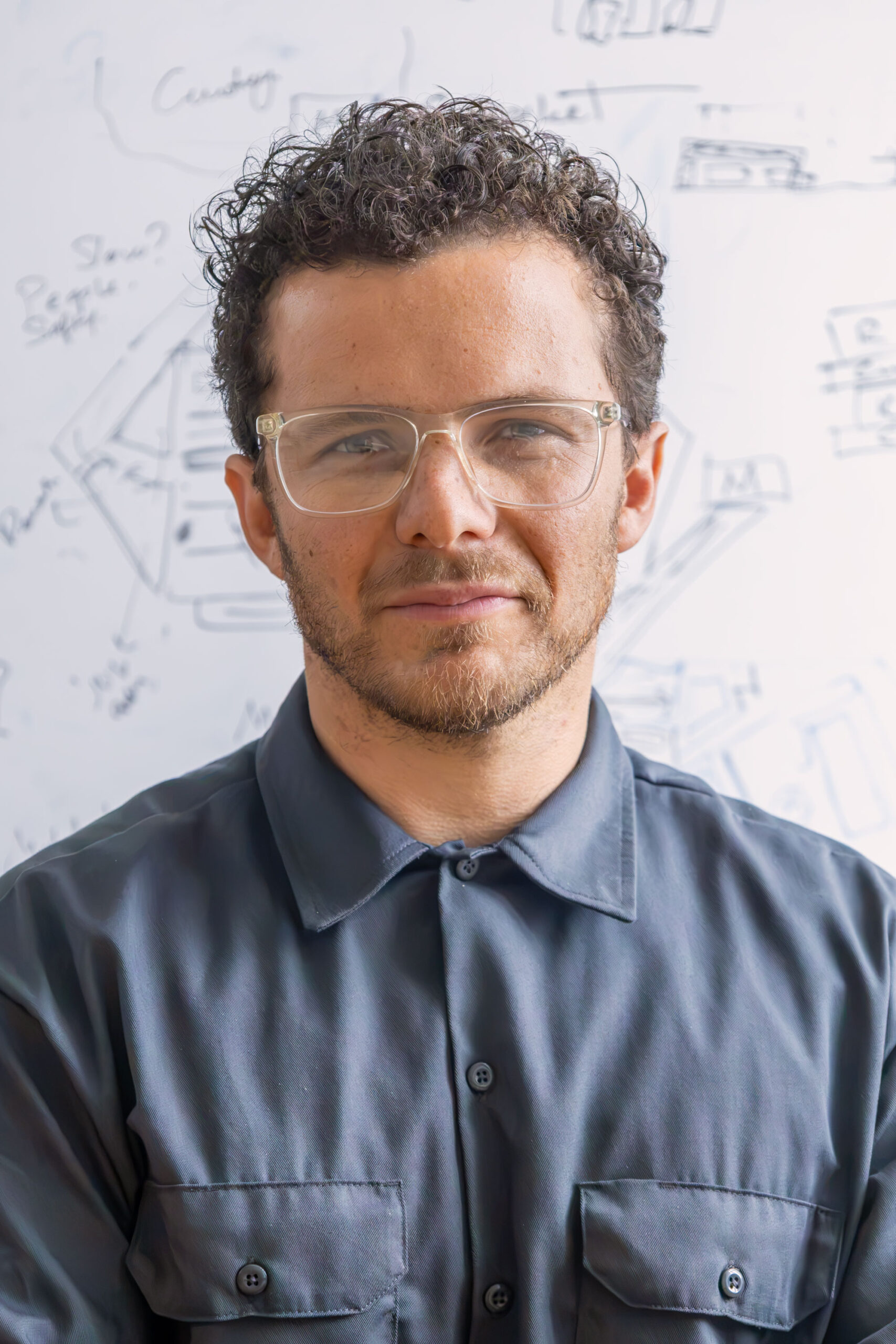A portrait of a man with short curly brown hair, clear rectangular glasses, and a short beard, wearing a grey button-down shirt with chest pockets. He is looking directly at the camera, standing in front of a whiteboard covered in faint sketches and handwritten notes.